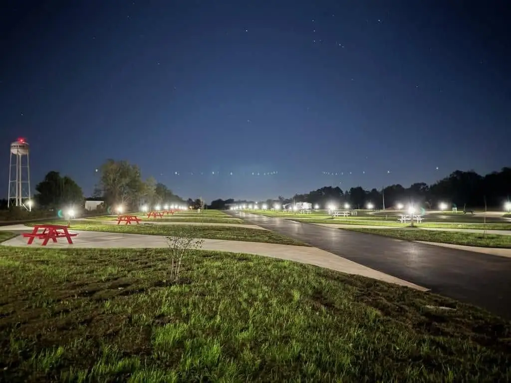 Nighttime view of an illuminated RV park with paved sites, red picnic tables on concrete pads, and a distant water