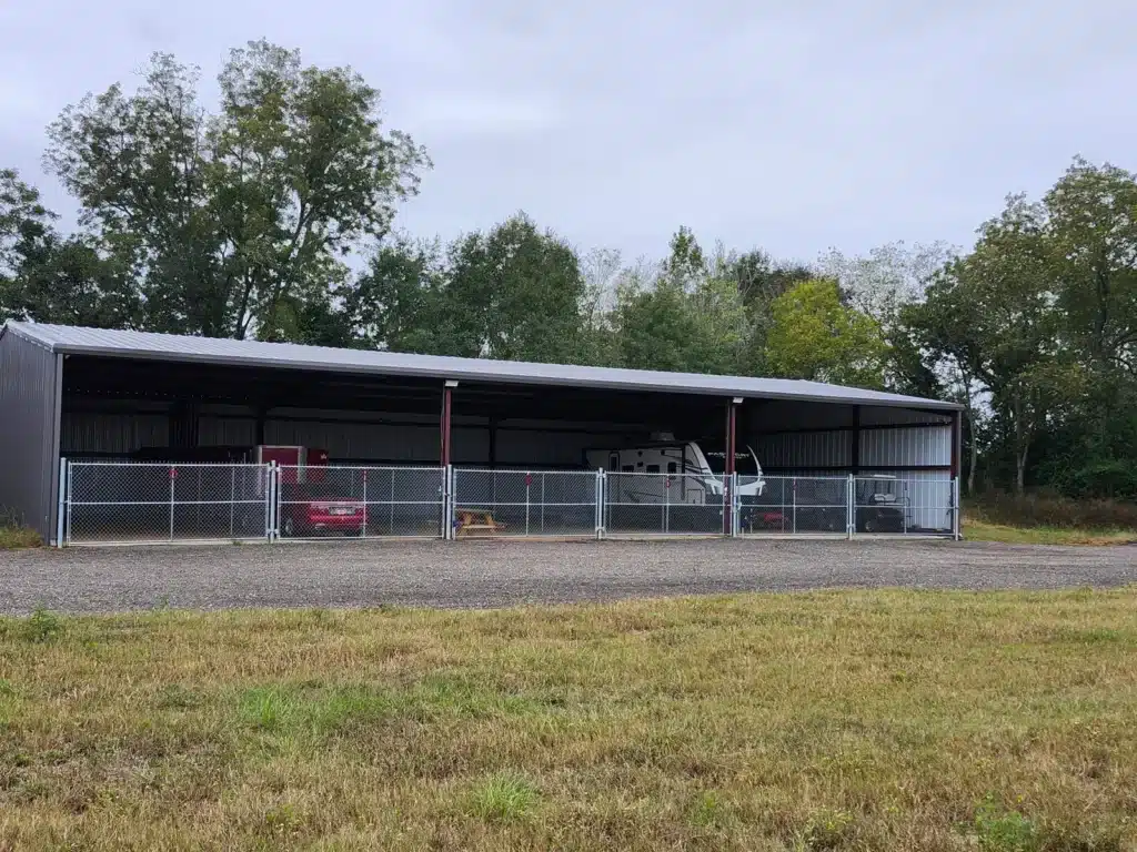 Long open-sided metal covered storage building behind a chain-link fence, sheltering a white travel trailer, red car