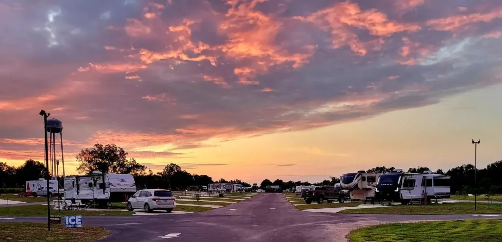 Vivid pink sunset over paved RV park road lined with travel trailers, parked cars, water tower, and an ICE sign.