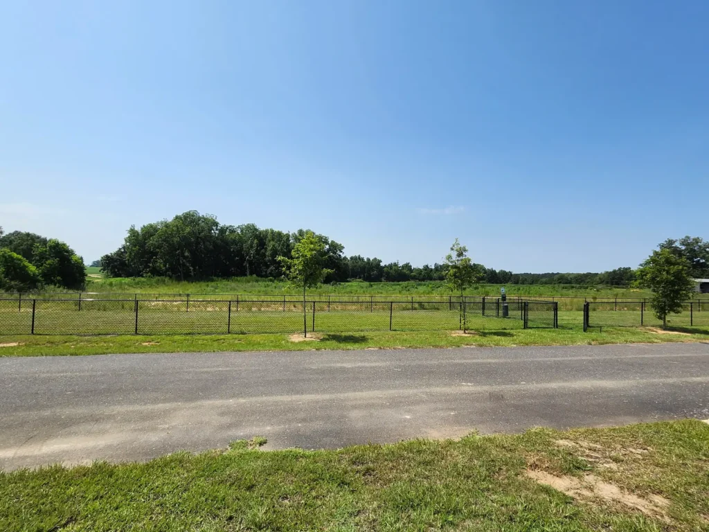 Paved road running past a black chain-link fence enclosing a mowed grassy field with young trees and distant treeline.